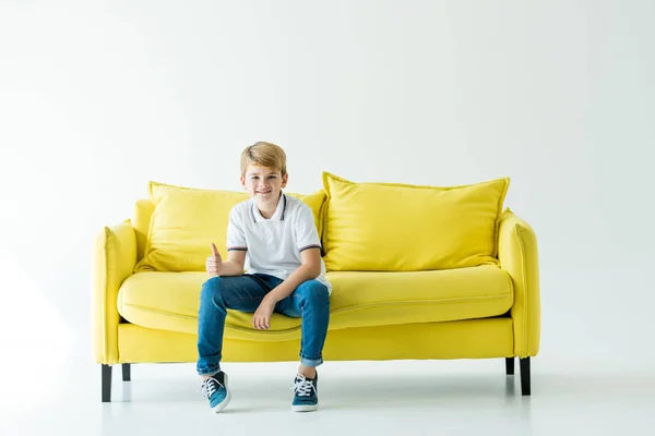 Sorrindo menino adorável sentado no sofá amarelo e mostrando o polegar para cima no branco — Fotografia de Stock