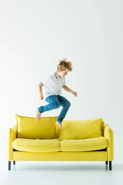 Entzückender Junge in lässiger Kleidung springt auf gelbem Sofa auf weißem — Stockfoto