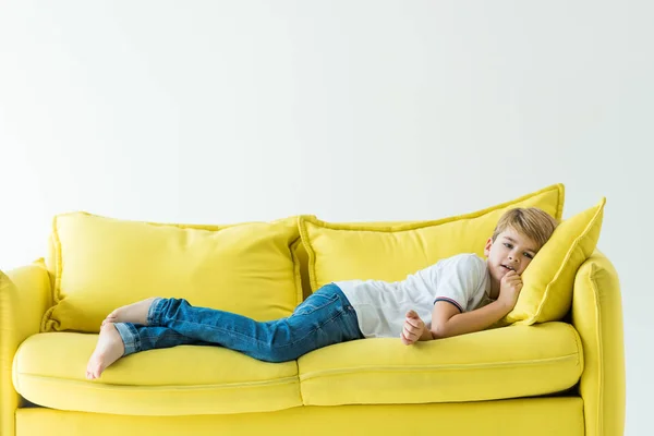 Menino adorável deitado em roupas casuais no sofá amarelo isolado no branco — Fotografia de Stock