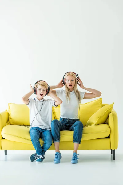 Schockierte Mutter und Sohn hören Musik mit Kopfhörern und berühren Köpfe auf gelbem Sofa auf weißem Grund — Stockfoto