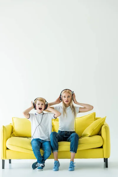 Conmocionados madre e hijo escuchando música con auriculares en sofá amarillo en blanco - foto de stock