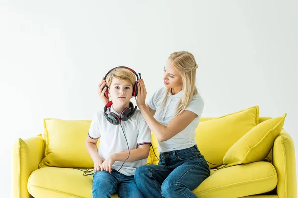 Mãe usando fones de ouvido no filho no sofá amarelo isolado no branco — Fotografia de Stock