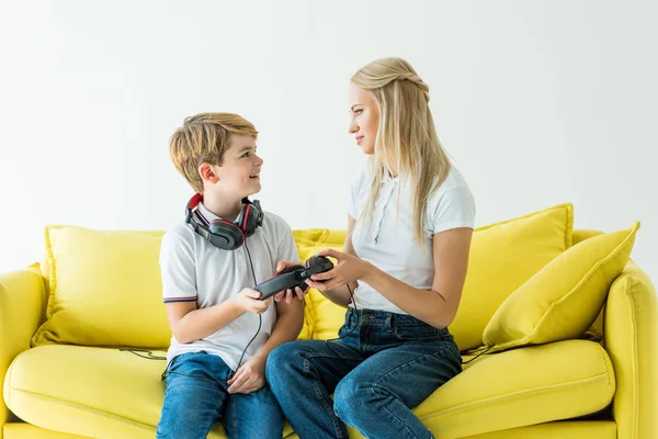 Glücklicher Sohn gibt Mutter Kopfhörer auf gelbem Sofa isoliert auf weißem — Stockfoto