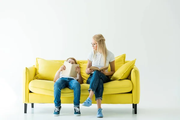 Mãe segurando livro e olhando para filho lendo livro no sofá amarelo no branco — Fotografia de Stock