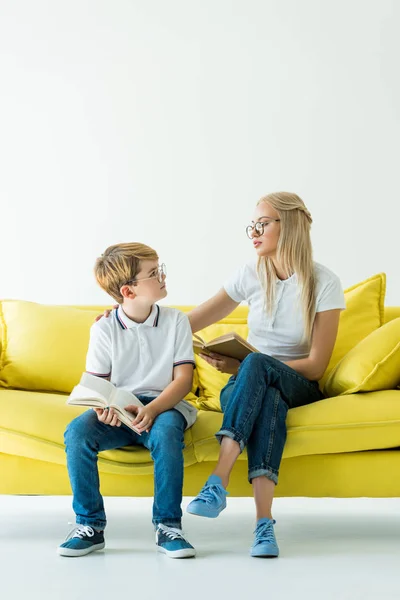 Mère et fils dans des lunettes tenant des livres et se regardant sur le canapé jaune — Photo de stock