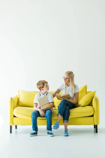 Madre e hijo sosteniendo libros y mirándose en un sofá amarillo - foto de stock