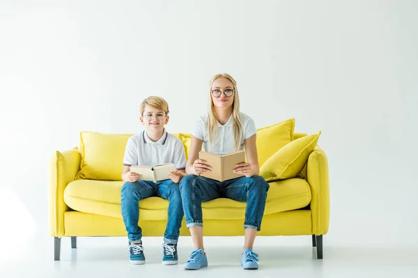 Smiling mother and son in glasses holding books and looking at camera on yellow sofa — Stock Photo