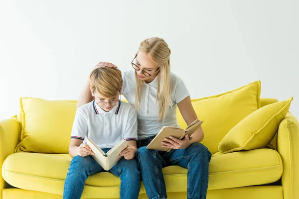 Mãe abraçando filho enquanto ele lendo livro no sofá amarelo isolado no branco — Fotografia de Stock