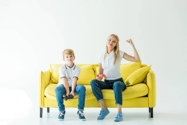 Feliz madre mostrando sí gesto después de ganar el videojuego en el sofá amarillo en blanco - foto de stock