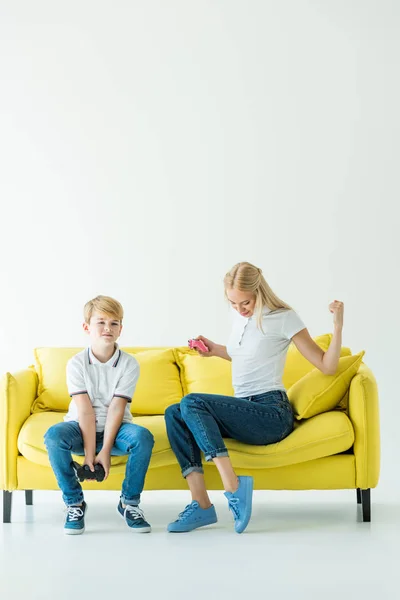 Мать показывает да жест после победы в видеоигре, сын сидит расстроенный на желтом диване на белом — стоковое фото
