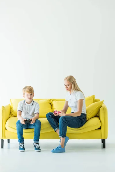 Mother and son sitting with gamepads on yellow sofa on white — Stock Photo