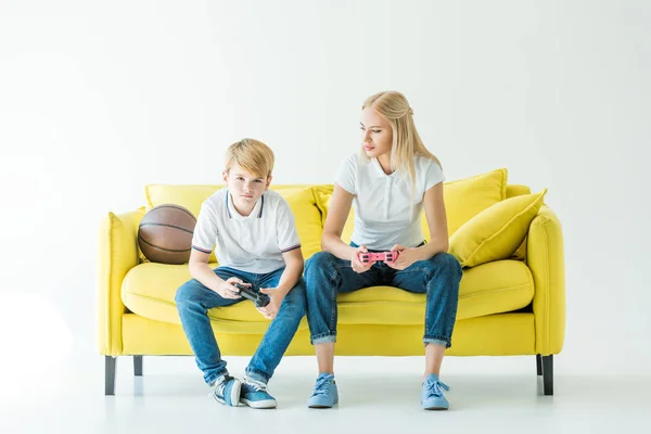 Madre mirando hijo mientras juega videojuego con mandos, pelota de baloncesto en sofá amarillo - foto de stock