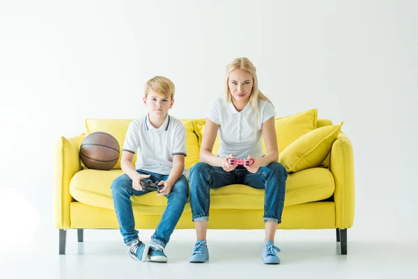 Seria madre e hijo jugando videojuego, pelota de baloncesto en sofá amarillo - foto de stock