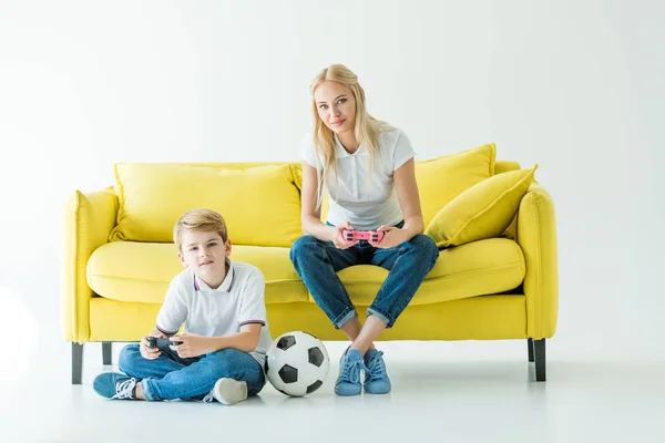 Madre e hijo jugando videojuego en sofá amarillo en blanco, pelota de fútbol en el suelo - foto de stock