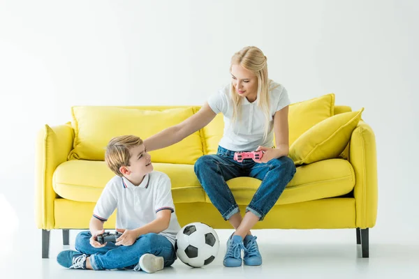 Mãe palming filho enquanto joga vídeo game no sofá amarelo no branco, bola de futebol no chão — Fotografia de Stock