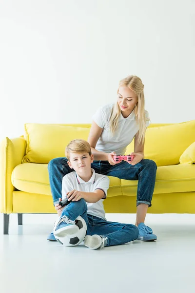Mãe olhando para o filho enquanto joga videogame no sofá amarelo no branco, bola de futebol no chão — Fotografia de Stock