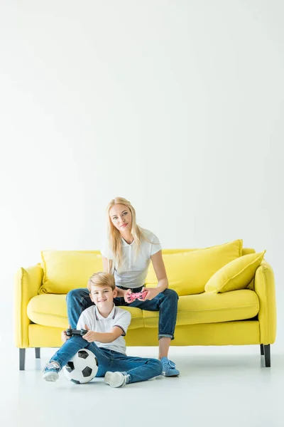 Heureux mère et fils jouer jeu vidéo sur canapé jaune sur blanc, ballon de football sur le sol — Photo de stock