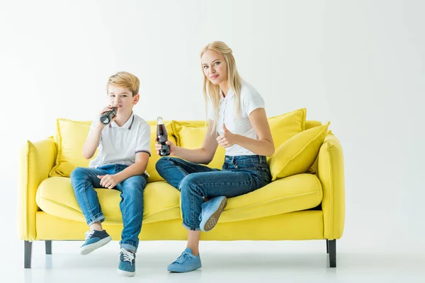 Madre mostrando el pulgar hacia arriba y el hijo bebiendo refresco en el sofá amarillo en blanco - foto de stock