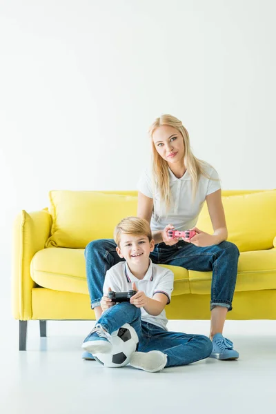 Sorrindo mãe e filho jogando videogame no sofá amarelo no branco, bola de futebol no chão — Fotografia de Stock