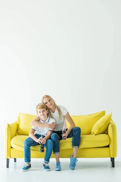 Mãe abraçando filho no sofá amarelo e eles segurando refrigerante em garrafas de vidro — Fotografia de Stock