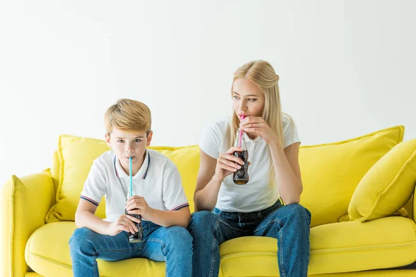 Mãe e filho bebendo refrigerante com palhas no sofá amarelo isolado em branco — Fotografia de Stock