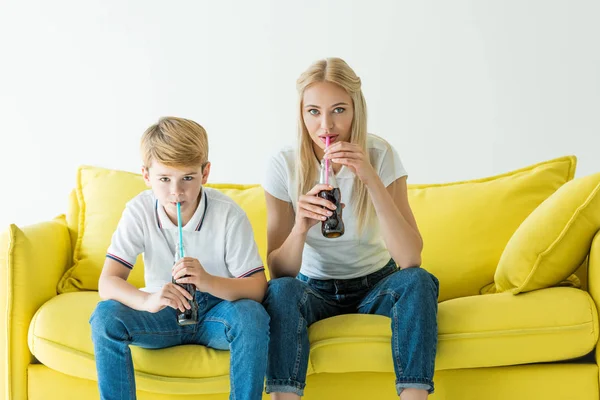 Mãe e filho bebendo refrigerante saboroso com palhas no sofá amarelo isolado no branco — Fotografia de Stock