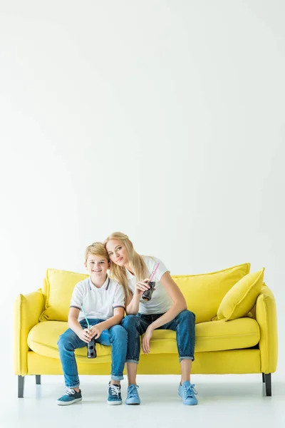 Mãe sorridente e filho segurando garrafas de refrigerante e sentado no sofá amarelo — Fotografia de Stock