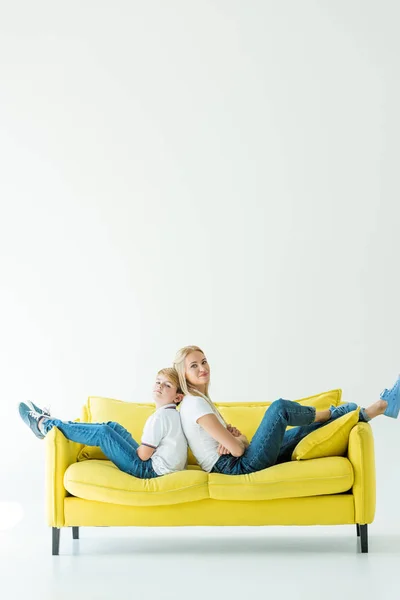 Madre e hijo sentados con los brazos cruzados en el sofá amarillo y mirando a la cámara en blanco - foto de stock