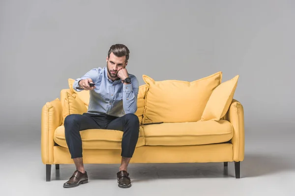 Bored man with remote control watching TV and sitting on yellow sofa on grey — Stock Photo