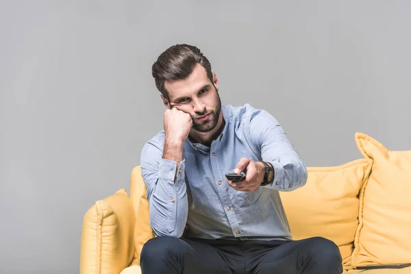 Schöner gelangweilter Mann mit Fernbedienung vor dem Fernseher auf gelbem Sofa auf grau — Stockfoto