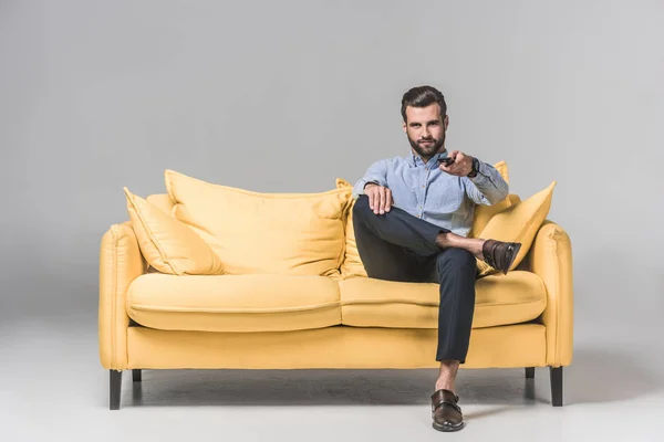 Bel homme souriant avec télécommande regardant la télévision et assis sur un canapé jaune sur gris — Photo de stock
