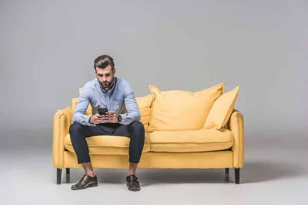 Elegant bored man using smartphone while sitting on yellow sofa on grey — Stock Photo