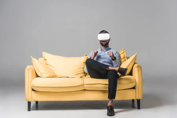 Мужчина использует гарнитуру виртуальной реальности и сидит на желтом диване на сером — стоковое фото