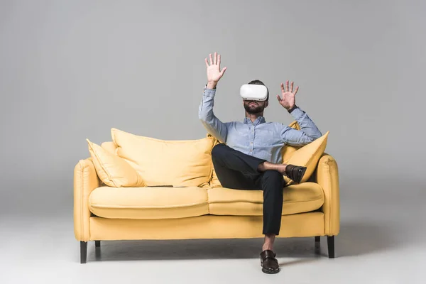 Uomo barbuto gesticolando e utilizzando auricolare realtà virtuale e seduto sul divano giallo sul grigio — Foto stock
