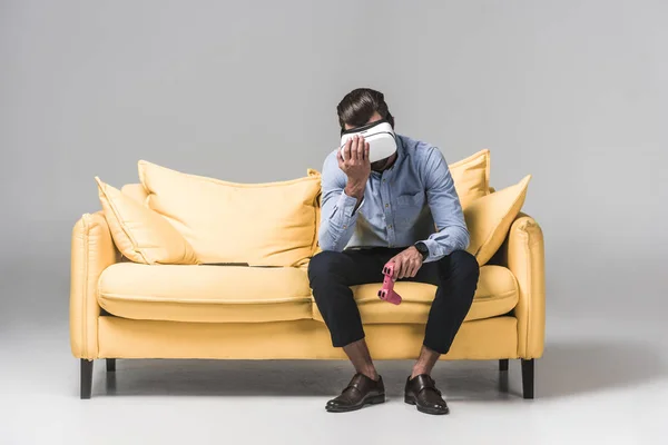 Расстроенный человек потерялся в видеоигре с джойстиком и гарнитурой виртуальной реальности на желтом диване на сером — стоковое фото