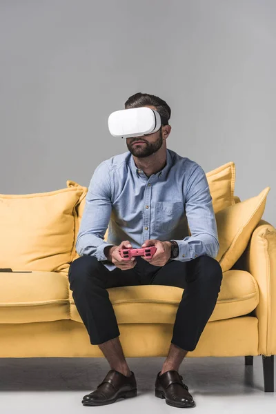 Чоловік грає у відеогру з джойстиком і гарнітурою vr на жовтому дивані на сірому — стокове фото