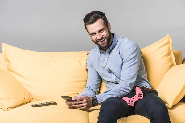 Homem sorridente usando smartphone e sentado no sofá amarelo com joystick e controle remoto em cinza — Fotografia de Stock