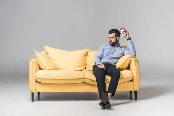Homme ennuyé dans l'usure formelle tenant café pour aller et assis sur un canapé jaune sur gris — Photo de stock