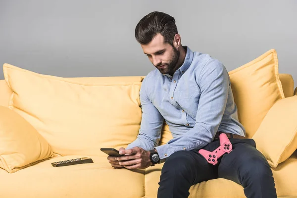 Hombre guapo usando teléfono inteligente y sentado en un sofá amarillo con joystick y control remoto en gris - foto de stock