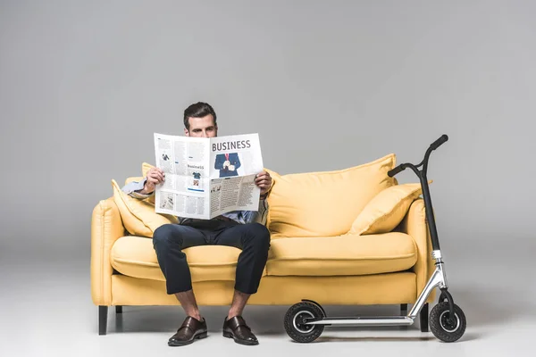 Serious man reading business newspaper while sitting on yellow sofa with scooter on grey — Stock Photo