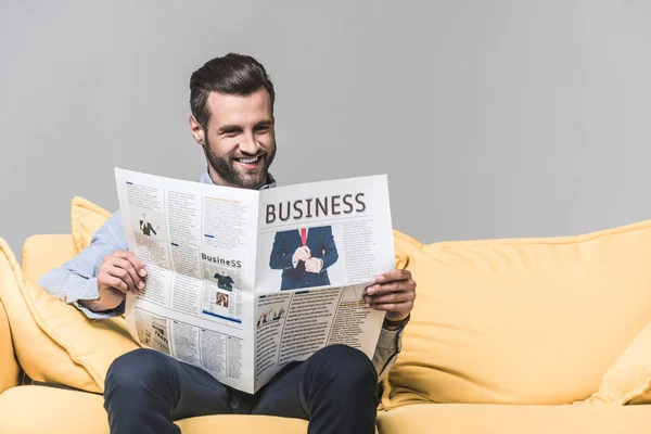 Homem barbudo sorridente lendo jornal de negócios enquanto sentado no sofá, no cinza — Fotografia de Stock