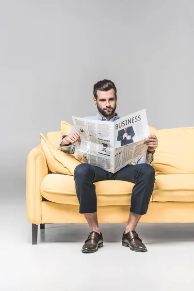 Élégant homme en tenue formelle lecture journal d'affaires tout en étant assis sur un canapé jaune, sur gris — Photo de stock