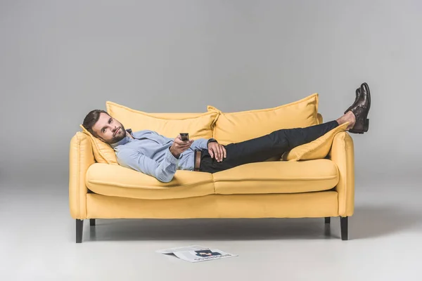 Bärtiger Mann mit Fernbedienung vor dem Fernseher und auf Sofa mit Zeitung auf dem Boden liegend, auf grau — Stockfoto
