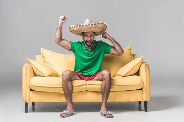 Bell'uomo in sombrero messicano che grida e festeggia il trionfo sul divano giallo su grigio — Foto stock