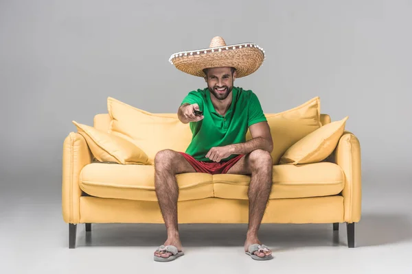 Smiling bearded man in mexican sombrero watching TV with remote control on yellow sofa on grey — Stock Photo