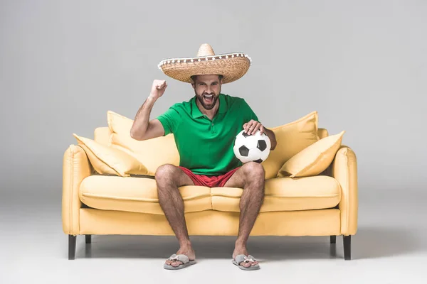Веселий чоловік в мексиканському сомбреро дивиться футбольний матч, сидячи на жовтому дивані з м'ячем на сірому — стокове фото