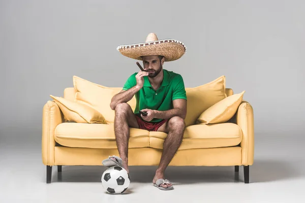 Serious man in mexican sombrero watching soccer match while sitting on yellow sofa with cigar, ball and remote control on grey — Stock Photo