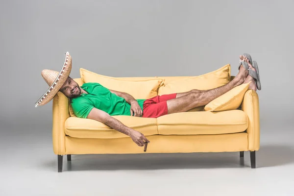 Happy man in mexican sombrero resting on yellow sofa with cigar on grey — Stock Photo