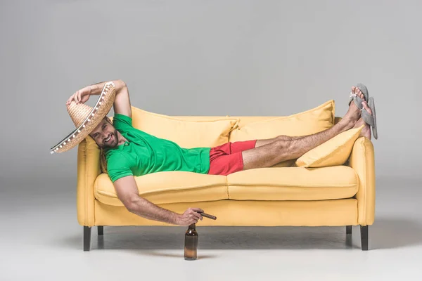 Homem sorridente no sombrero mexicano relaxando no sofá amarelo com garrafa de cerveja e charuto no cinza — Fotografia de Stock