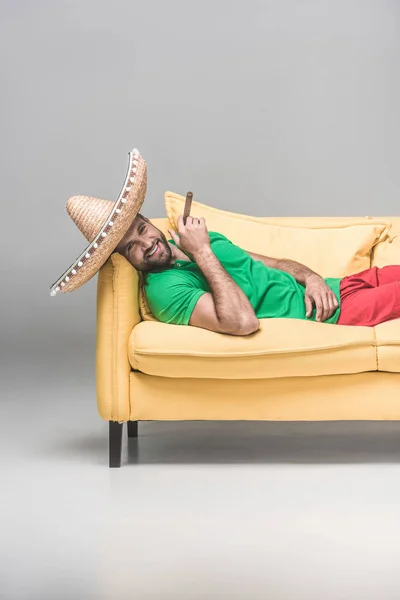 Красивый веселый мужчина в мексиканском сомбреро с сигарой, лежащей на желтом диване на сером — стоковое фото